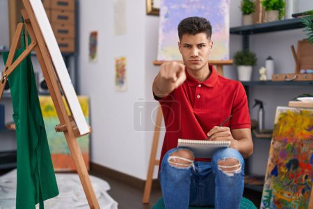 Téléchargez les photos : Jeune homme hispanique dessinant sur un carnet d'art pointant du doigt la caméra et vers vous, geste confiant et sérieux - en image libre de droit