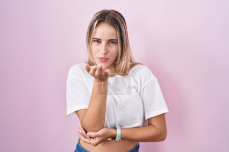 Foto de Young blonde woman standing over pink background looking at the camera blowing a kiss with hand on air being lovely and sexy. love expression. - Imagen libre de derechos