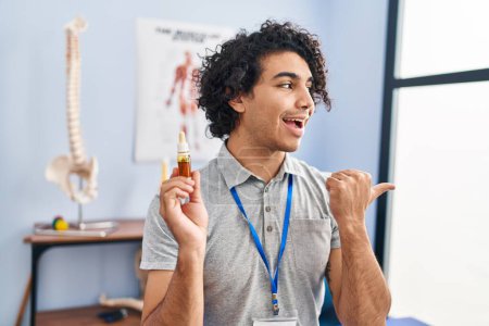 Photo for Hispanic man with curly hair holding cbd oil pointing thumb up to the side smiling happy with open mouth - Royalty Free Image