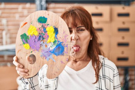 Foto de Mujer hispana de mediana edad sosteniendo paleta cubriendo la cara sacando la lengua feliz con expresión divertida. - Imagen libre de derechos