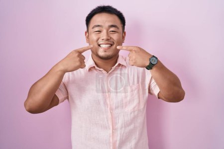 Téléchargez les photos : Jeune homme chinois debout sur fond rose souriant gai montrant et pointant avec les doigts dents et la bouche. concept de santé dentaire. - en image libre de droit