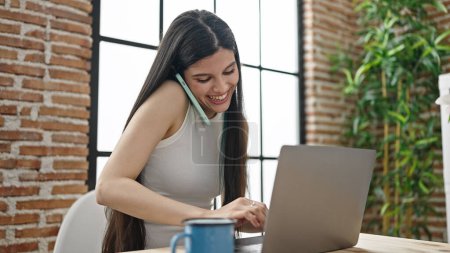 Photo for Young beautiful hispanic woman talking on smartphone using laptop at dinning room - Royalty Free Image