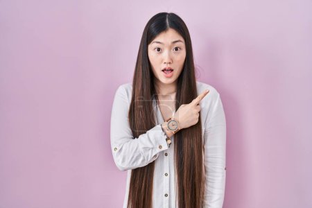 Photo for Chinese young woman standing over pink background surprised pointing with finger to the side, open mouth amazed expression. - Royalty Free Image