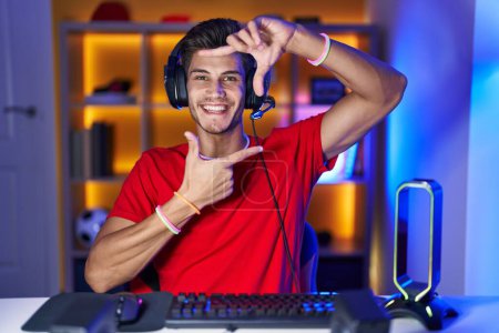 Foto de Joven hombre hispano jugando videojuegos sonriente haciendo marco con manos y dedos con cara feliz. concepto de creatividad y fotografía. - Imagen libre de derechos