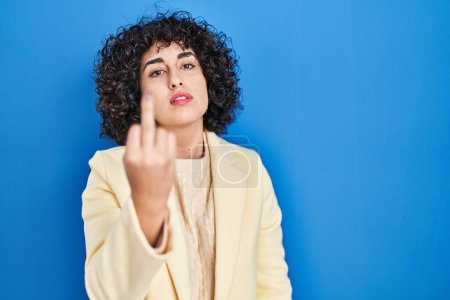 Photo for Young brunette woman with curly hair standing over blue background showing middle finger, impolite and rude fuck off expression - Royalty Free Image