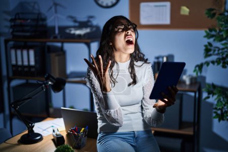 Photo for Young brazilian woman using touchpad at night working at the office crazy and mad shouting and yelling with aggressive expression and arms raised. frustration concept. - Royalty Free Image