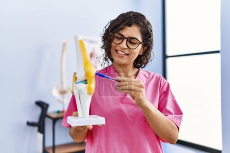 Téléchargez les photos : Young latin woman wearing physiotherapist uniform holding anatomical model of knee at clinic - en image libre de droit
