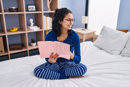 Foto de Joven mujer latina leyendo libro sentado en la cama en el dormitorio - Imagen libre de derechos