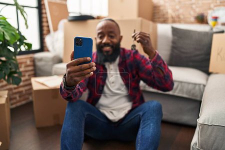 Téléchargez les photos : Jeune homme afro-américain faire selfie par le smartphone détenant la clé de la nouvelle maison à la nouvelle maison - en image libre de droit