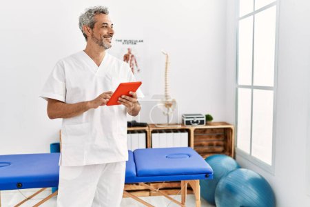 Foto de Fisioterapeuta hombre de pelo gris de mediana edad sonriendo confiado usando touchpad en la clínica de rehabilitación - Imagen libre de derechos
