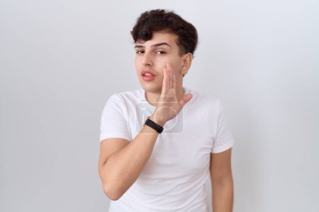 Téléchargez les photos : Jeune homme non binaire portant casual t-shirt blanc main sur la bouche racontant rumeur secrète, chuchotant conversation malicieuse - en image libre de droit