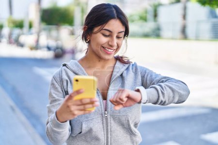 Téléchargez les photos : Young hispanic woman wearing sportswear looking stopwatch using smartphone at street - en image libre de droit
