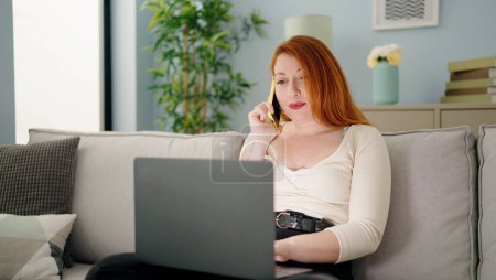 Foto de Mujer pelirroja joven usando el ordenador portátil y hablando en el teléfono inteligente en casa - Imagen libre de derechos