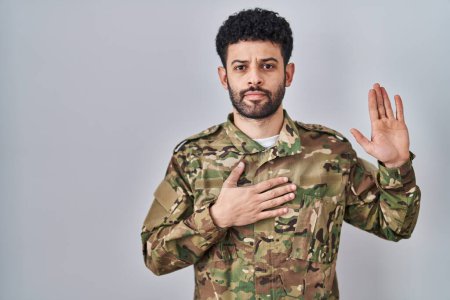 Téléchargez les photos : Homme arabe portant un uniforme de camouflage de l'armée jurant avec la main sur la poitrine et la paume ouverte, faisant un serment de fidélité - en image libre de droit