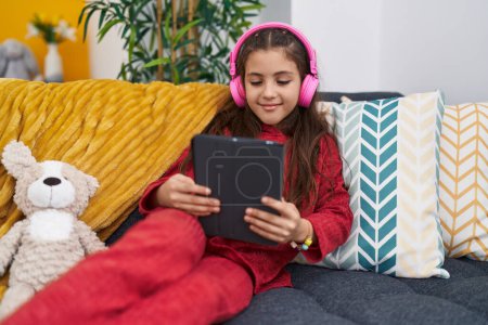 Photo for Adorable hispanic girl using touchpad and headphones sitting on sofa at home - Royalty Free Image
