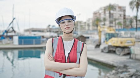 Téléchargez les photos : Jeune belle bâtisseur hispanique debout avec expression sérieuse et bras croisés geste au bord de la mer - en image libre de droit