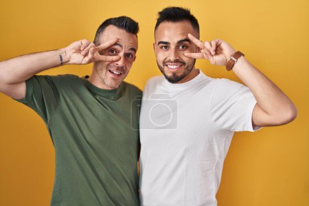 Téléchargez les photos : Couple homosexuel debout sur fond jaune faisant symbole de paix avec les doigts sur le visage, souriant gai montrant la victoire - en image libre de droit