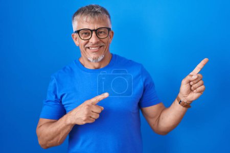 Photo for Hispanic man with grey hair standing over blue background smiling and looking at the camera pointing with two hands and fingers to the side. - Royalty Free Image