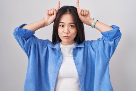 Photo for Young chinese woman standing over white background doing funny gesture with finger over head as bull horns - Royalty Free Image