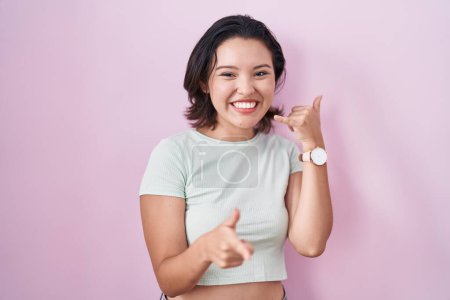 Téléchargez les photos : Jeune femme hispanique debout sur fond rose souriant faire parler au téléphone geste et vous pointant du doigt. Appelle-moi. - en image libre de droit