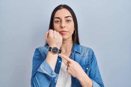 Photo for Hispanic woman standing over blue background in hurry pointing to watch time, impatience, looking at the camera with relaxed expression - Royalty Free Image
