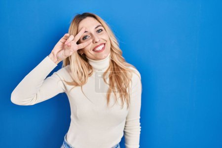 Foto de Young caucasian woman standing over blue background doing peace symbol with fingers over face, smiling cheerful showing victory - Imagen libre de derechos