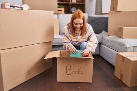 Photo for Young caucasian woman smiling confident unpacking books cardboard box at new home - Royalty Free Image