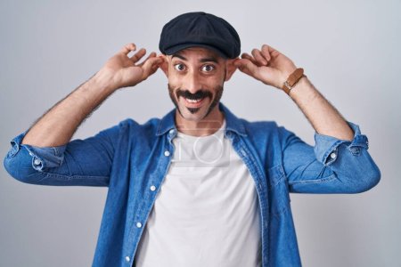 Téléchargez les photos : Hispanic man with beard standing over isolated background smiling pulling ears with fingers, funny gesture. audition problem - en image libre de droit
