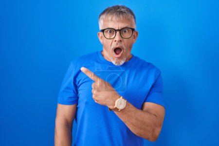 Photo for Hispanic man with grey hair standing over blue background surprised pointing with finger to the side, open mouth amazed expression. - Royalty Free Image
