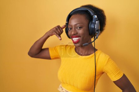 Téléchargez les photos : Femme africaine avec les cheveux bouclés debout sur fond jaune portant des écouteurs souriant faire geste de téléphone avec la main et les doigts comme parler au téléphone. communiquer les concepts. - en image libre de droit