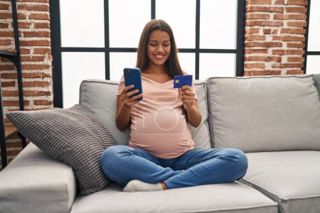 Téléchargez les photos : Jeune femme latine enceinte avec smartphone et carte de crédit à la maison - en image libre de droit