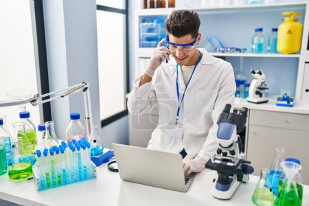 Photo for Young hispanic man wearing scientist uniform using laptop and talking on the smartphone at laboratory - Royalty Free Image