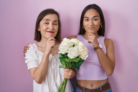 Foto de Madre e hija hispanas sosteniendo un ramo de flores blancas mirando confiadas a la cámara sonriendo con los brazos cruzados y la mano levantada en la barbilla. pensar positivamente. - Imagen libre de derechos