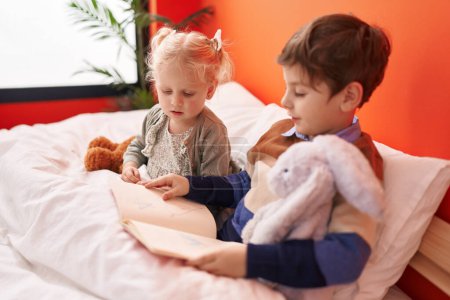 Foto de Adorable niño y niña leyendo libro sentado en la cama en el dormitorio - Imagen libre de derechos