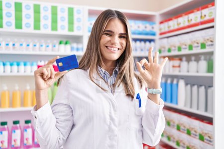 Foto de Mujer joven hispana trabajando en farmacia sosteniendo tarjeta de crédito haciendo signo de bien con los dedos, sonriendo gesto amistoso excelente símbolo - Imagen libre de derechos