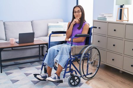 Photo for Young hispanic woman sitting on wheelchair at home looking confident at the camera smiling with crossed arms and hand raised on chin. thinking positive. - Royalty Free Image