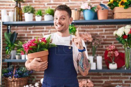 Foto de Joven hispano trabajando en floristería sosteniendo maceta sonriendo con una idea o pregunta señalando con el dedo con cara feliz, número uno - Imagen libre de derechos