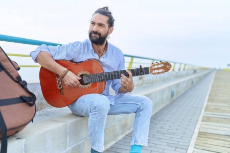 Téléchargez les photos : Young hispanic man musician playing classical guitar sitting on bench at seaside - en image libre de droit