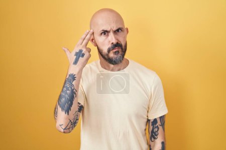 Foto de Hombre hispano con tatuajes de pie sobre fondo amarillo disparando y matándose apuntando de la mano y los dedos a la cabeza como arma, gesto suicida. - Imagen libre de derechos