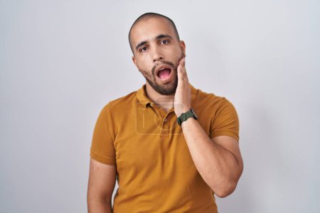 Foto de Hispanic man with beard standing over white background touching mouth with hand with painful expression because of toothache or dental illness on teeth. dentist - Imagen libre de derechos