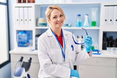 Téléchargez les photos : Jeune femme blonde scientifique souriante confiante tenant des lunettes au laboratoire - en image libre de droit