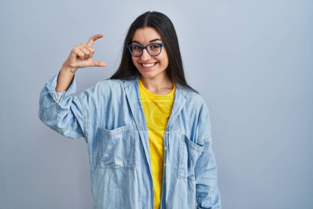 Téléchargez les photos : Jeune femme hispanique debout sur fond bleu geste souriant et confiant avec la main faisant signe de petite taille avec les doigts regardant et la caméra. concept de mesure. - en image libre de droit