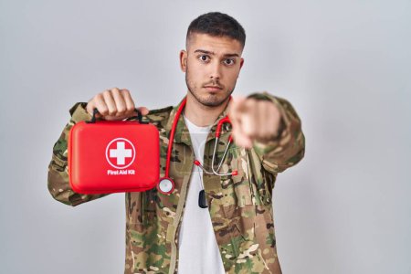 Photo for Young hispanic doctor wearing camouflage army uniform holding first aid kit pointing with finger to the camera and to you, confident gesture looking serious - Royalty Free Image