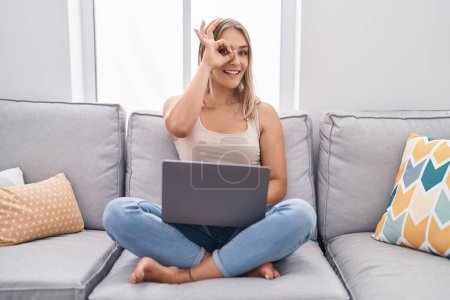Foto de Rubia mujer caucásica usando el ordenador portátil en casa sentado en el sofá sonriendo feliz haciendo signo bien con la mano en el ojo mirando a través de los dedos - Imagen libre de derechos