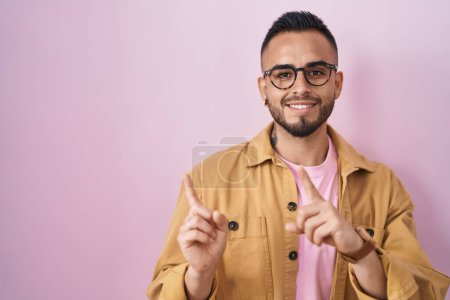 Foto de Young hispanic man standing over pink background smiling and looking at the camera pointing with two hands and fingers to the side. - Imagen libre de derechos