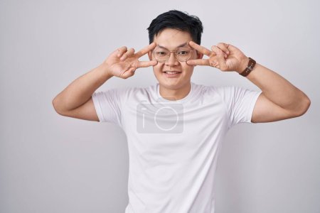 Téléchargez les photos : Jeune homme asiatique debout sur fond blanc faisant symbole de paix avec les doigts sur le visage, souriant joyeux montrant la victoire - en image libre de droit