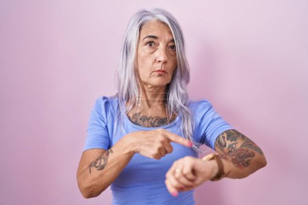 Téléchargez les photos : Femme du moyen âge avec des tatouages debout sur fond rose à la hâte pointant vers regarder le temps, l'impatience, bouleversé et en colère pour retard de délai - en image libre de droit