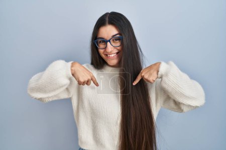 Foto de Mujer hispana joven vistiendo suéter casual sobre fondo azul mirando confiado con sonrisa en la cara, señalándose con los dedos orgullosos y felices. - Imagen libre de derechos