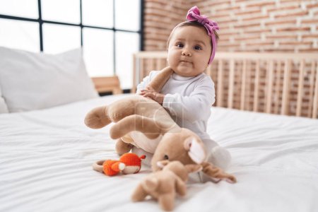 Téléchargez les photos : Adorable bébé hispanique assis sur le lit avec une expression détendue dans la chambre - en image libre de droit