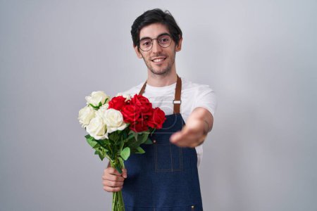 Photo for Young hispanic man holding bouquet of white and red roses smiling cheerful offering palm hand giving assistance and acceptance. - Royalty Free Image